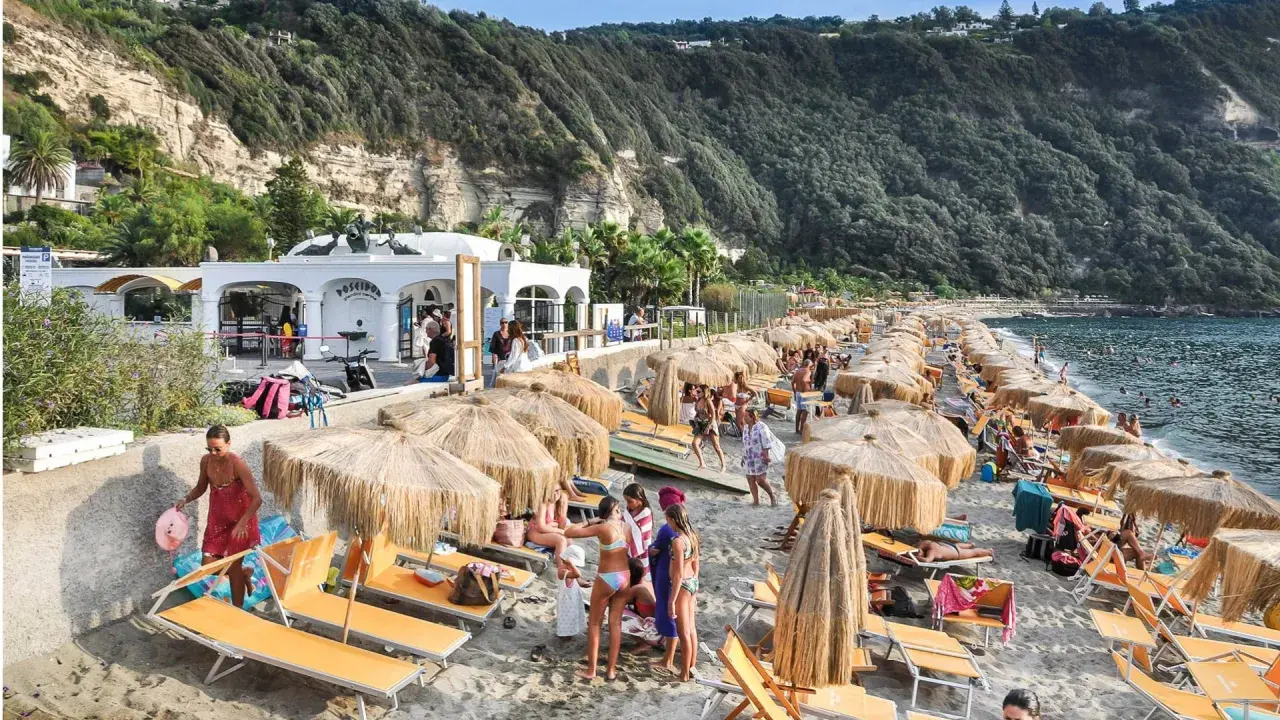 Spiaggia di Citara affollata, con ombrelloni di paglia, lettini arancioni e gente che si gode il mare. Per arrivare, segui le indicazioni per Forio.