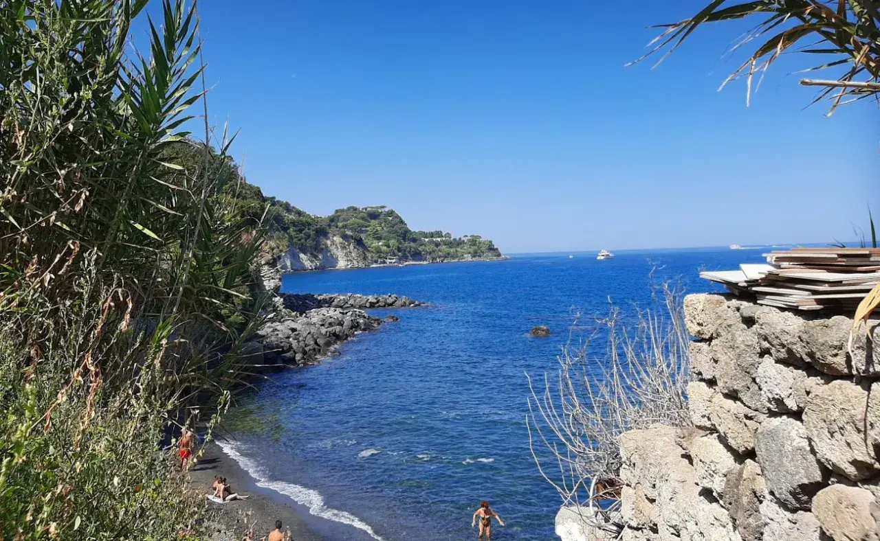 Spiaggia nera con gente che si gode il sole, scogli e il blu intenso del mare degli inglesi.