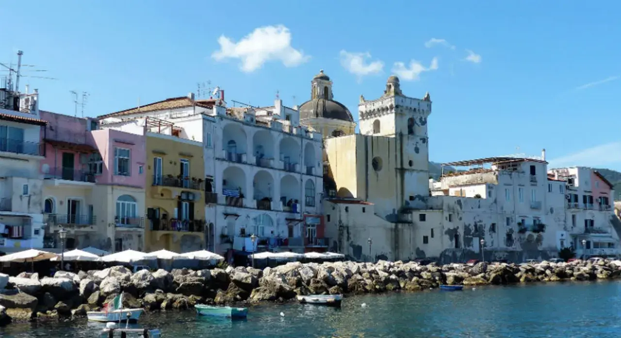 Colorate case affacciate sul mare, un porto con barche e rocce. Per sapere come arrivare ad Ischia in treno, si può prendere un treno per Napoli e poi un aliscafo.