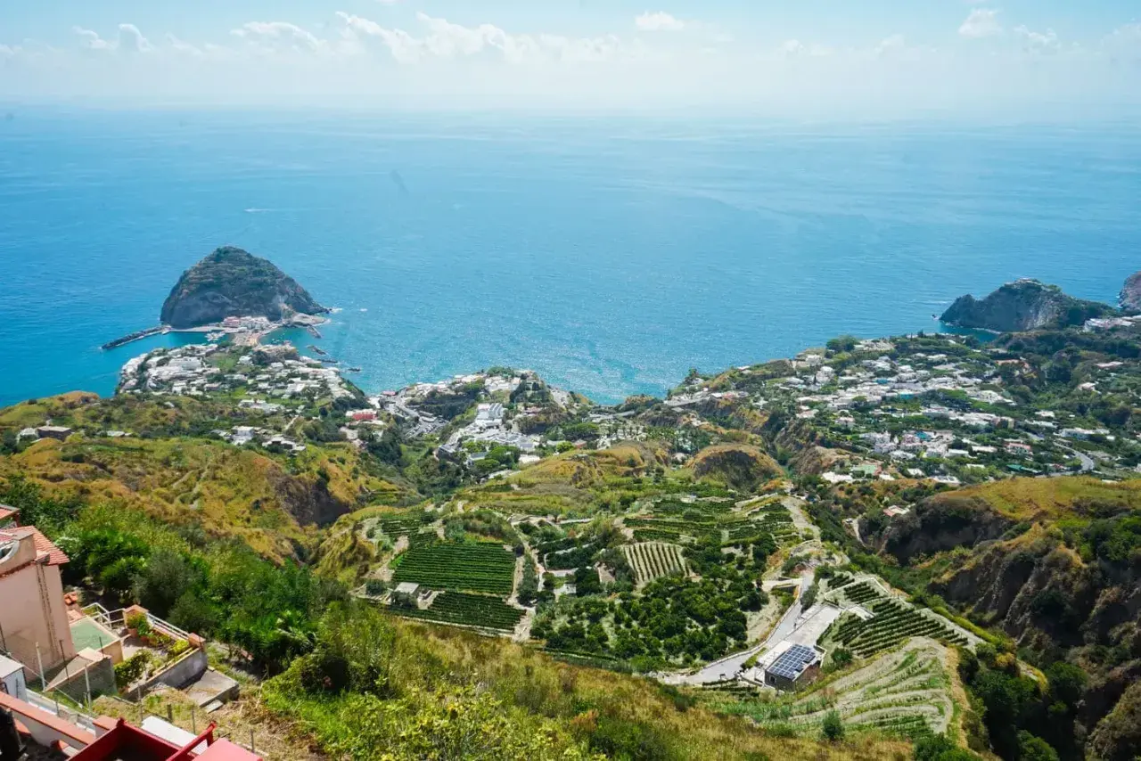 Panoramica mozzafiato di Ischia, con terrazzamenti verdi e il mare blu. Si intravede una casa museo, immersa in questo paesaggio incantevole.