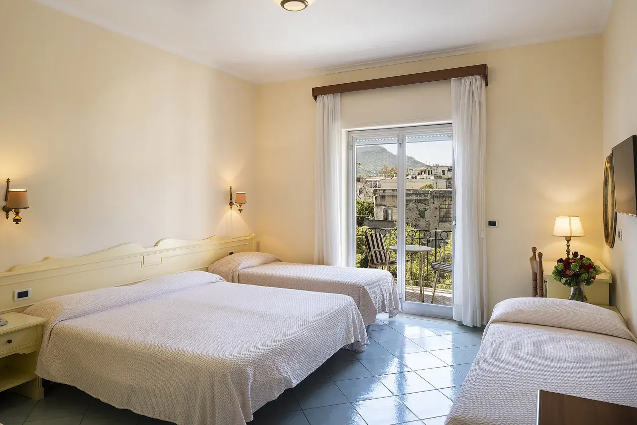 Camera con tre letti singoli, vista su un borgo di Alexander Ischia. Balcone con tavolo e sedie.