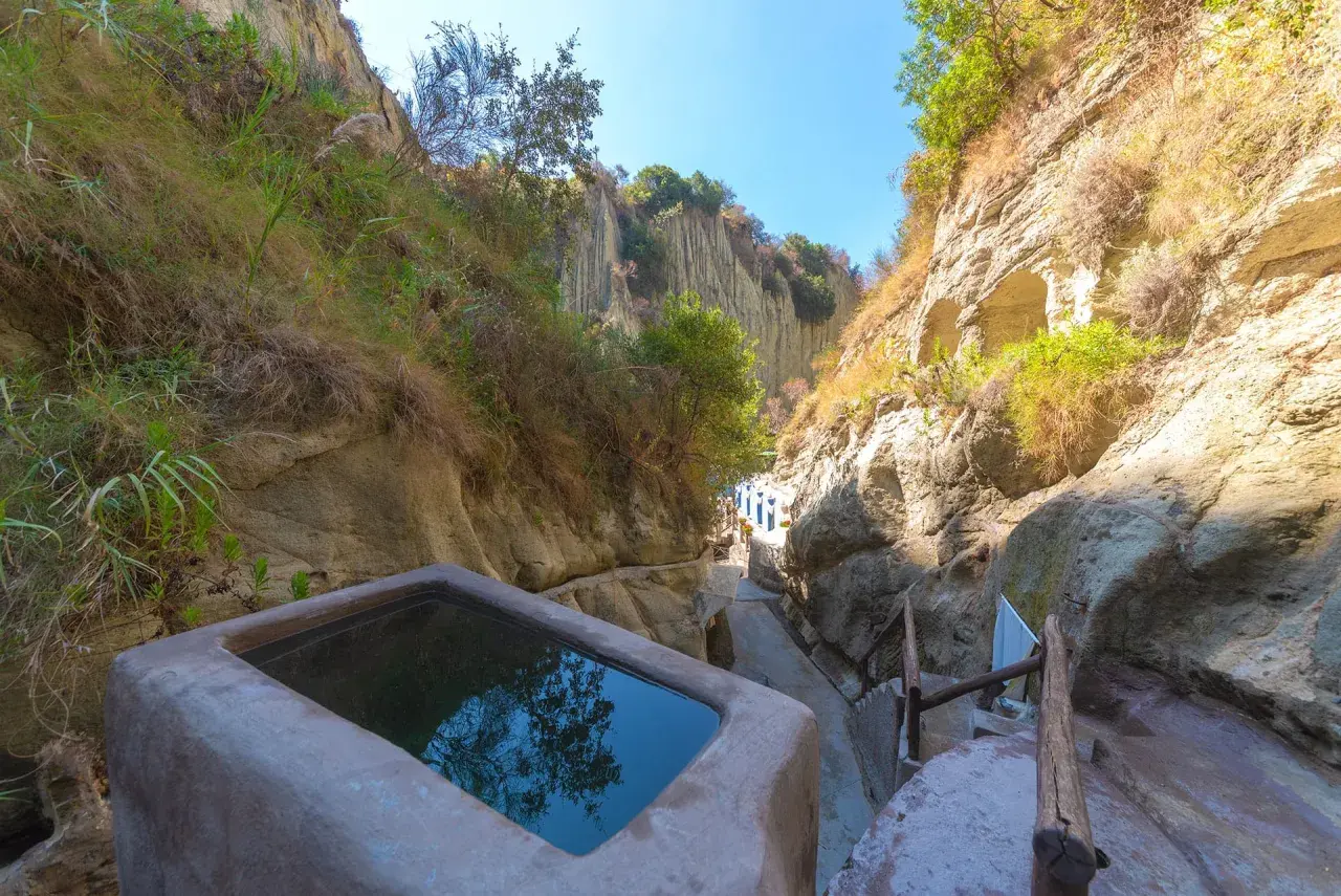 Piscina naturale scavata nella roccia, parte delle storiche terme di Cavascura, circondata da vegetazione e pareti rocciose.