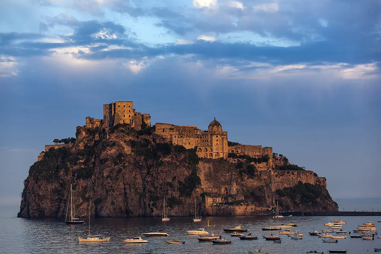 Il maestoso Castello Aragonese di Ischia si erge su uno scoglio roccioso, circondato da barche al tramonto.