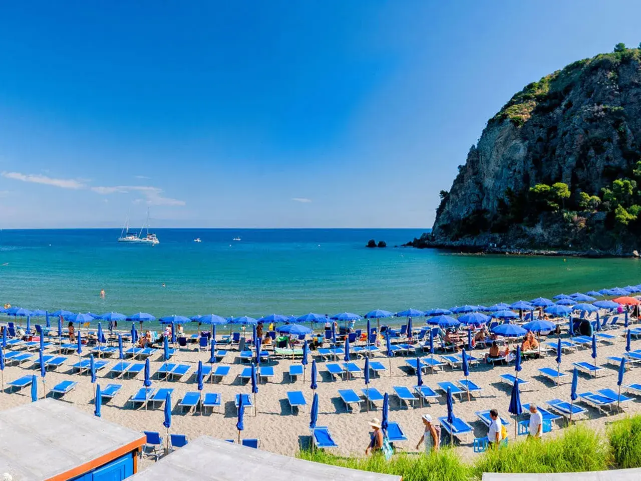 Spiaggia affollata con ombrelloni blu e lettini sulla riviera di San Montano. Mare azzurro e scogliera verde sullo sfondo.