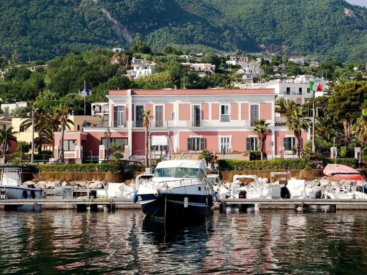 Il **hotel Svizzera Ischia** si affaccia sul porto con barche ormeggiate. Sullo sfondo, case arroccate su una collina verdeggiante.