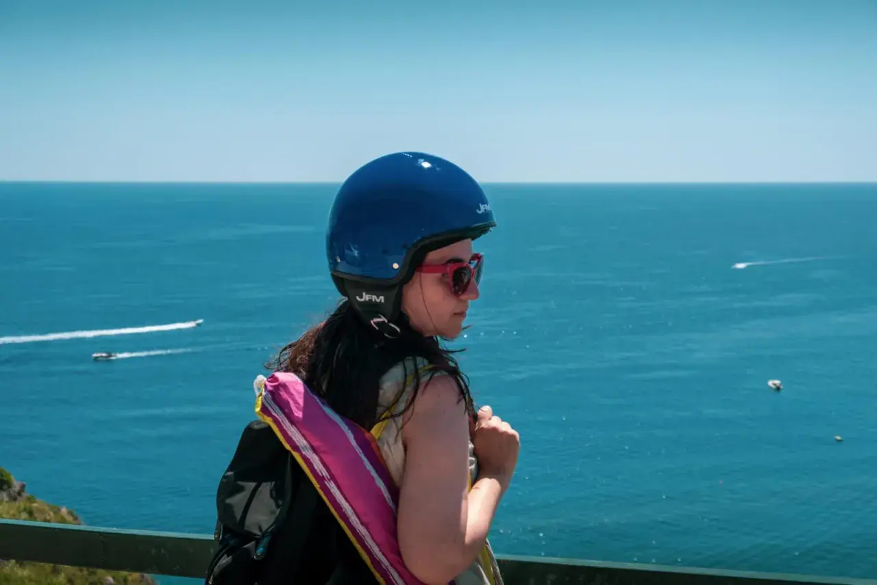 Ragazza con casco blu e zaino, guarda il mare. Pensa a come arrivare ad Ischia in auto, magari con uno scooter.