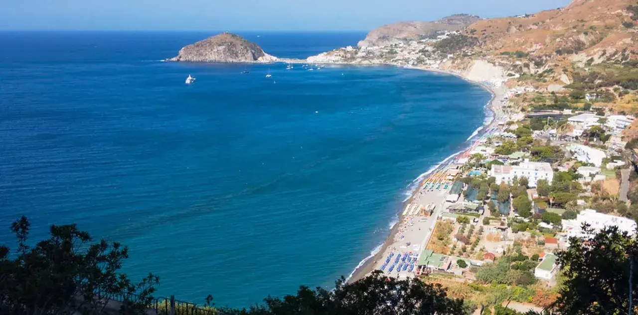 Vista panoramica della spiaggia dei Maronti con lidi attrezzati, ombrelloni colorati e il mare blu intenso. In lontananza, un isolotto e la costa frastagliata.
