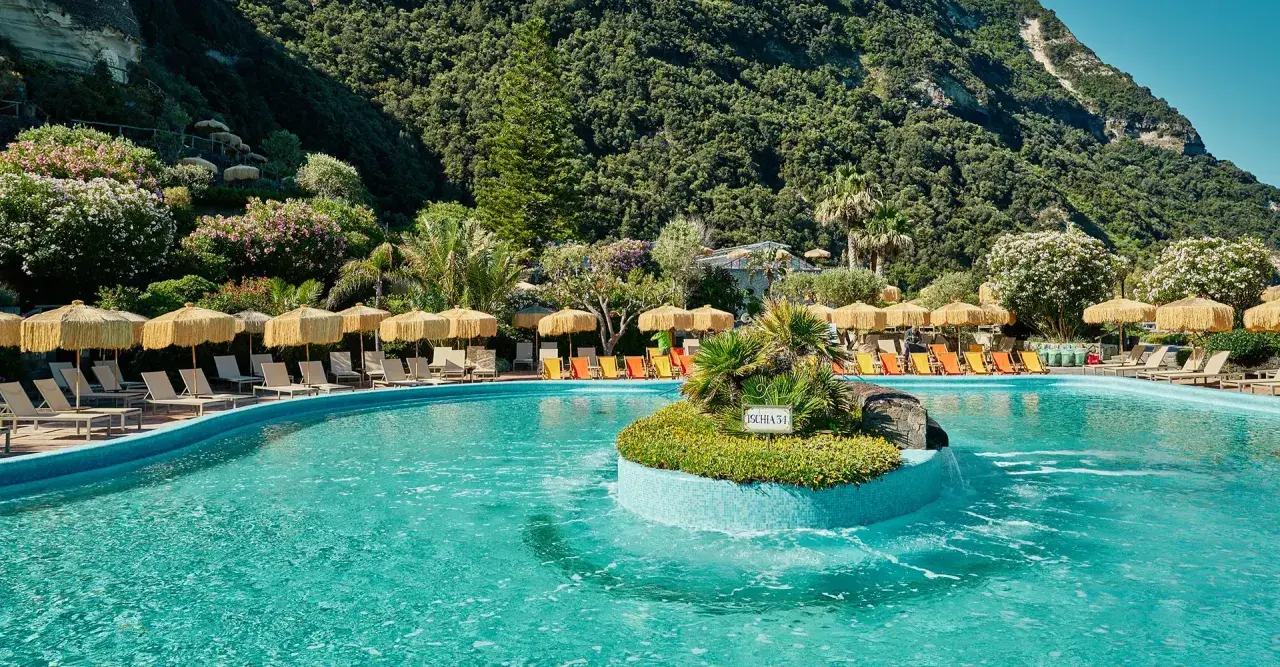 Piscina con lettini e ombrelloni a Ischia, immersa nel verde rigoglioso. Un'oasi di relax che ricorda la forza del **Tifeo** che dorme sotto l'isola.