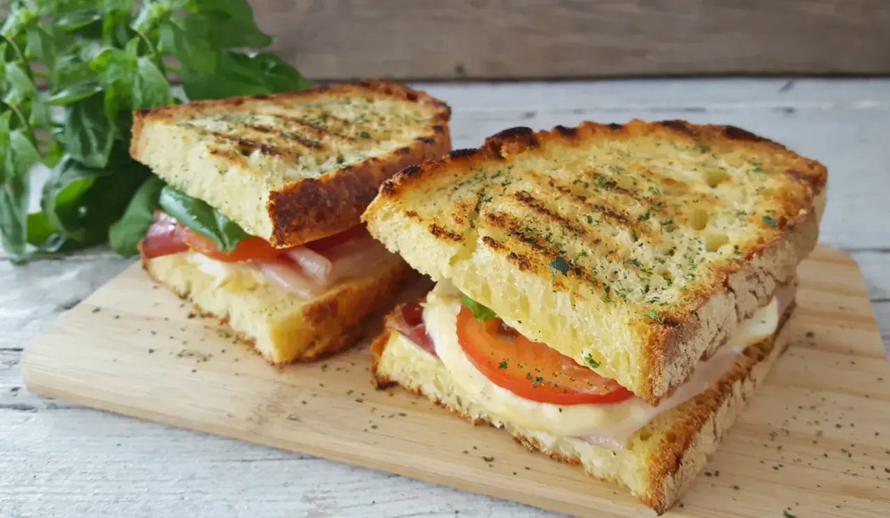 Due panini grigliati, una zingara ischitana, con pomodoro, mozzarella filante e prosciutto, su tagliere di legno.
