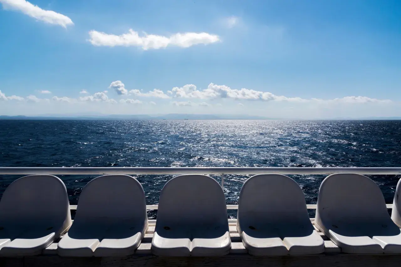 Sedili vuoti su un traghetto, con il mare blu scintillante e le montagne all'orizzonte. La differenza tra aliscafo e traghetto è evidente nella velocità e nel comfort.
