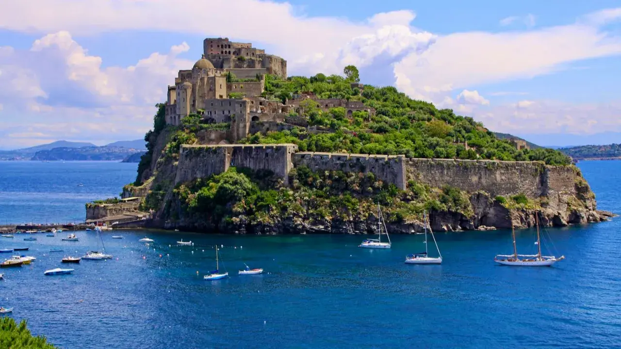 Ischia Ponte, con il maestoso Castello Aragonese che domina l'isola verdeggiante, circondato da un mare blu intenso e barche a vela.