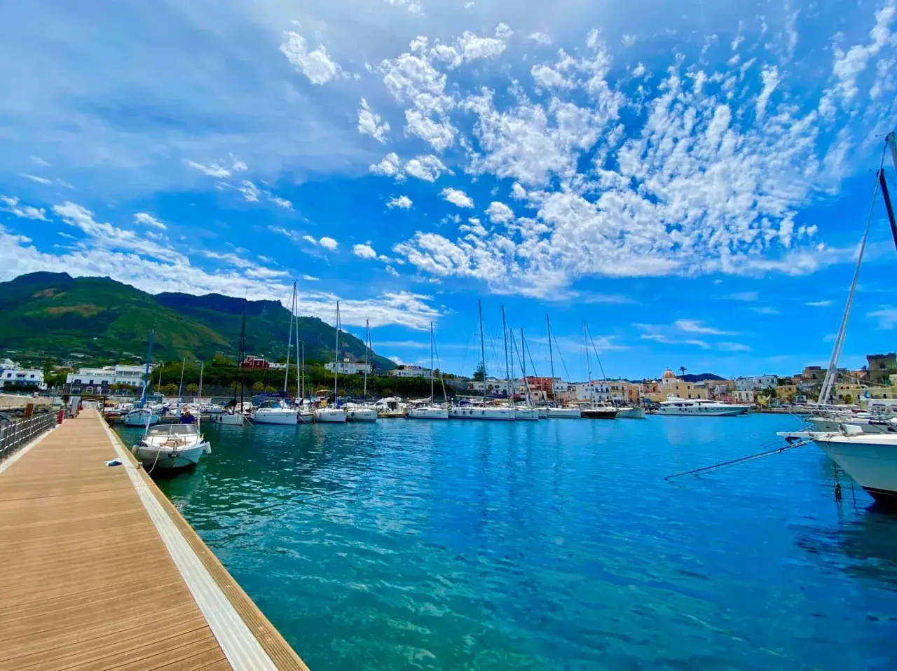 Porto di Forio, Ischia: barche a vela ormeggiate in un mare azzurro sotto un cielo con nuvole bianche. Per raggiungere Forio da Ischia Porto, si può prendere un autobus o un taxi.
