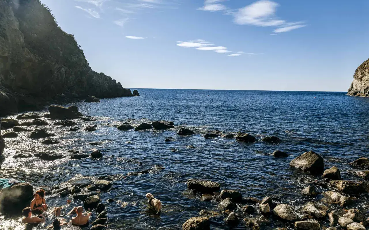Persone e un cane si godono le acque calde della **baia di Sorgeto**, tra scogli e rocce a picco sul mare blu.