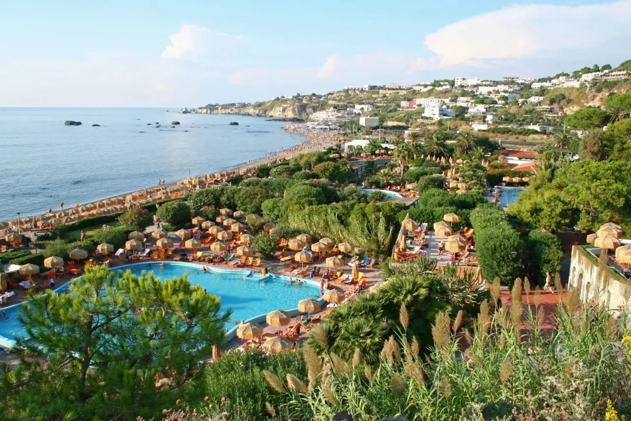 Vista panoramica di un resort a Ischia con piscine, lettini, ombrelloni e la spiaggia che si estende verso il mare.