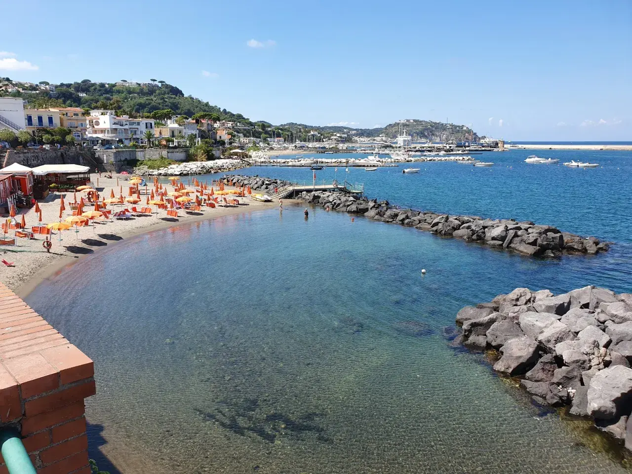 La spiaggia della marina è baciata dal sole, con ombrelloni arancioni e lettini pronti per accogliere i bagnanti. Il mare cristallino invita a un tuffo.