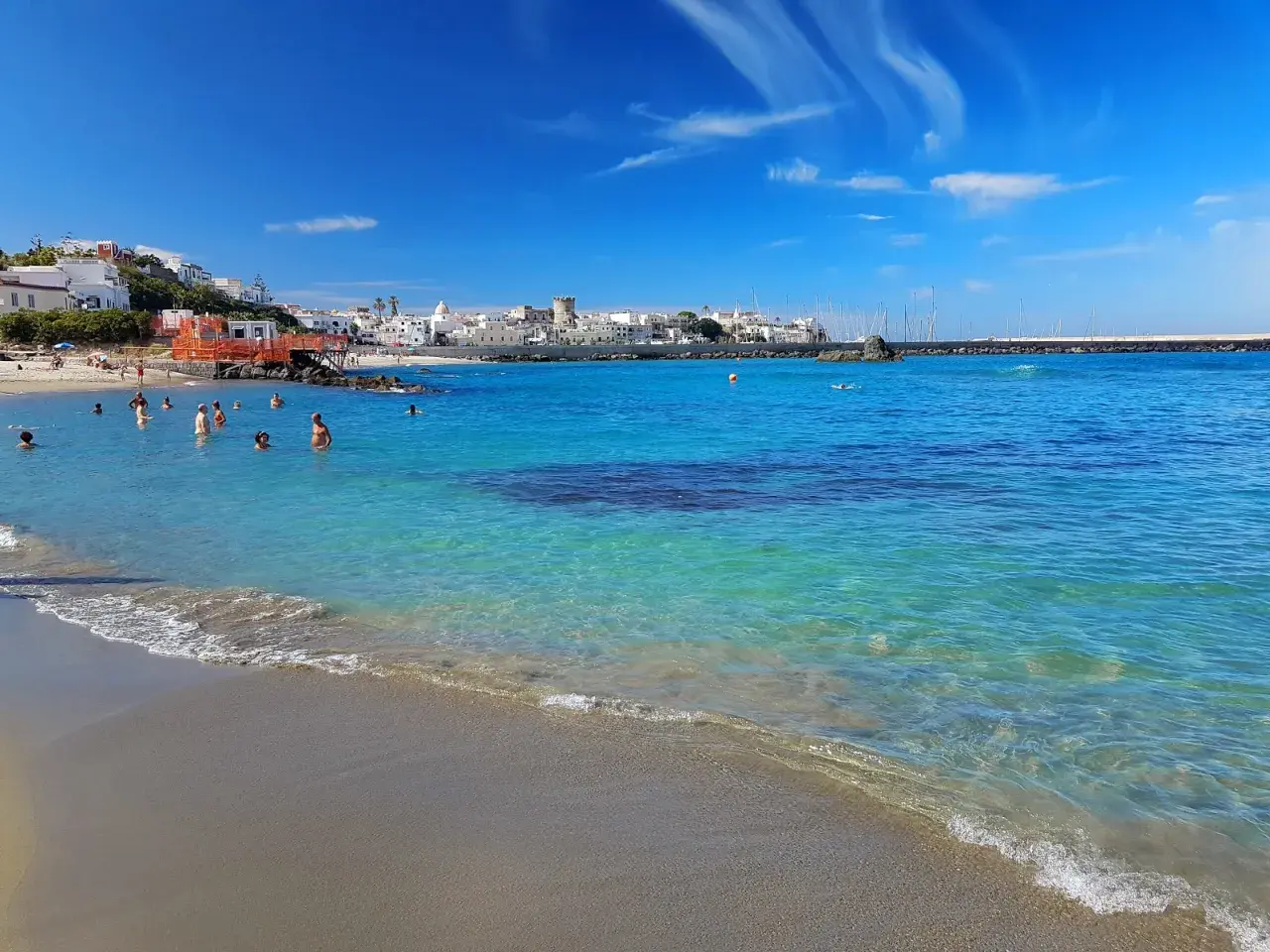 Persone nuotano nelle acque cristalline della spiaggia della Chiaia, con un borgo bianco sullo sfondo e un cielo azzurro con nuvole bianche.