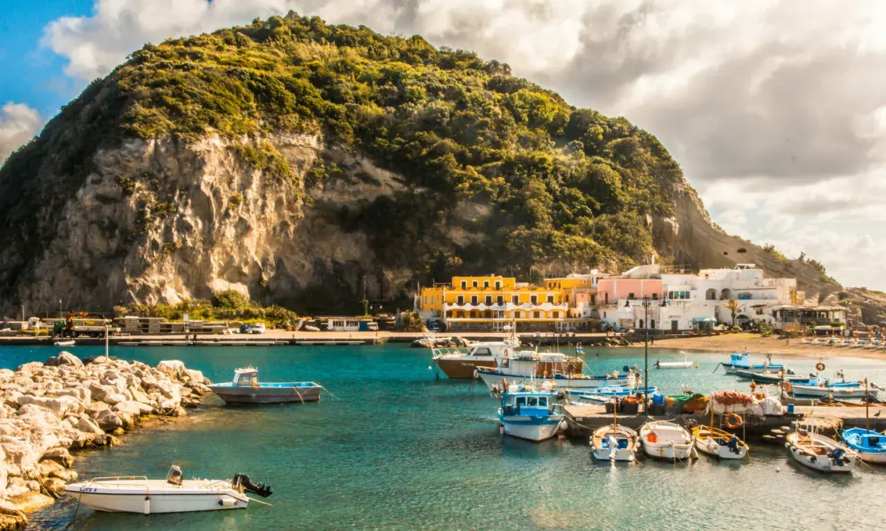 Un pittoresco ormeggio a Ischia, con barche colorate ormeggiate davanti a case bianche e gialle, sotto una collina verdeggiante.