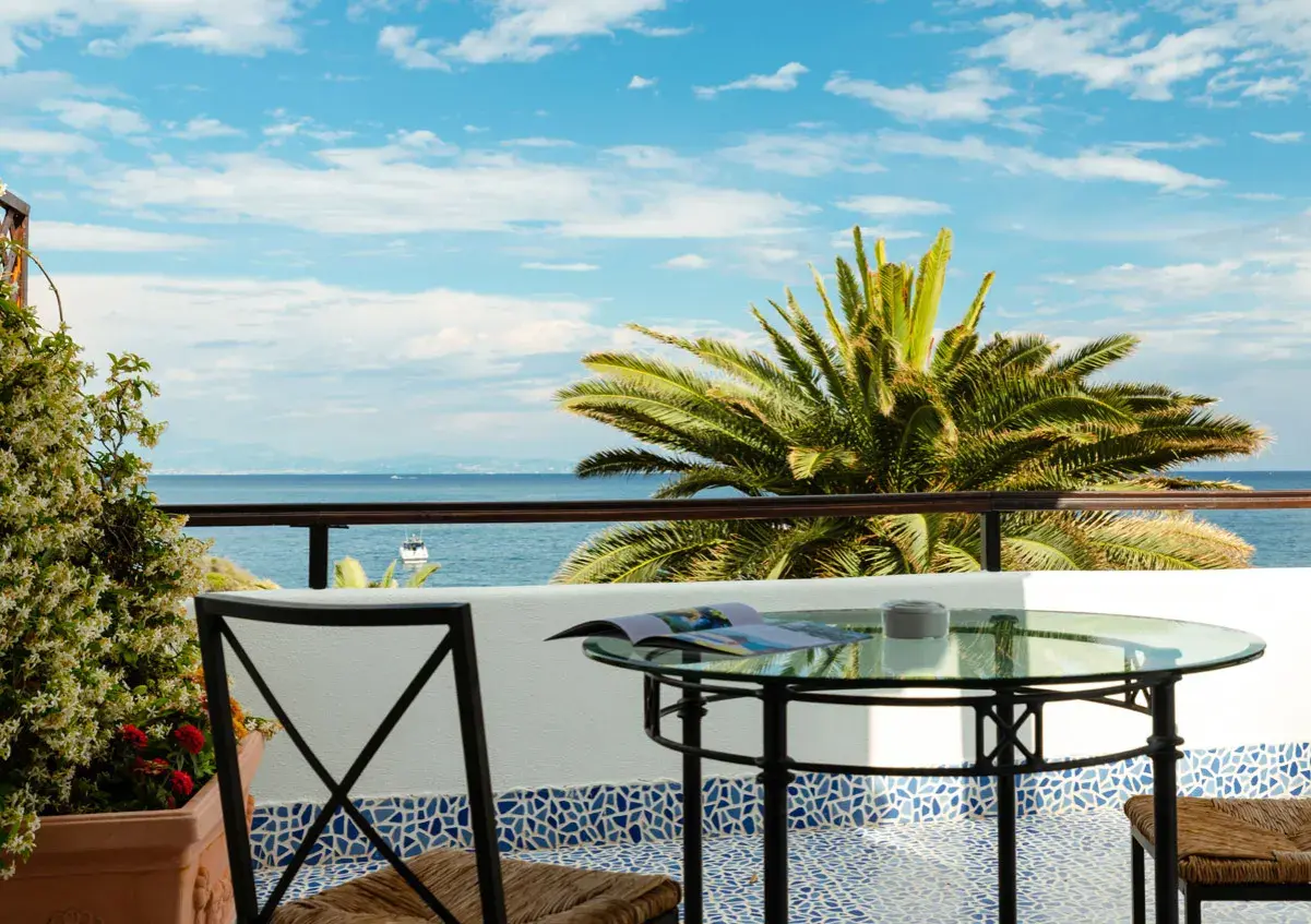 Vista mare da un beach club a Ischia: tavolino in vetro, sedia in ferro e palma rigogliosa sotto un cielo azzurro.
