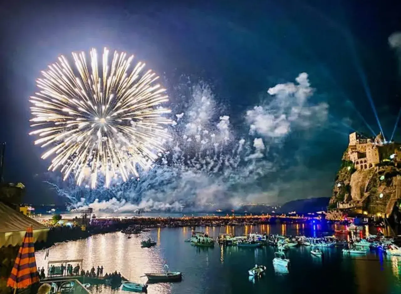 Fuochi d'artificio spettacolari illuminano il cielo notturno sopra un porto pieno di barche, con un castello arroccato su una scogliera. Un'immagine che ricorda le celebrazioni di Mario Mazzella.