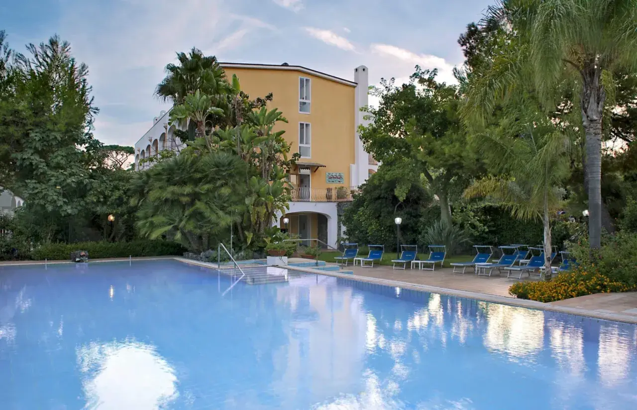 Piscina azzurra e lettini blu in un rigoglioso giardino tropicale, con un edificio giallo sullo sfondo, a San Giovanni, Ischia.