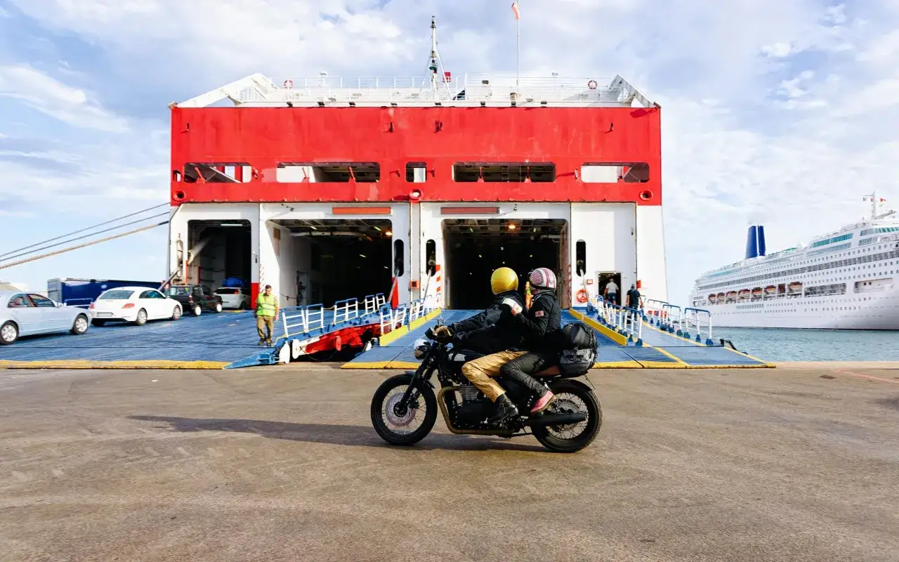 Coppia in moto sbarca da un traghetto, pronta per esplorare Ischia. Sullo sfondo, un'altra nave da crociera.