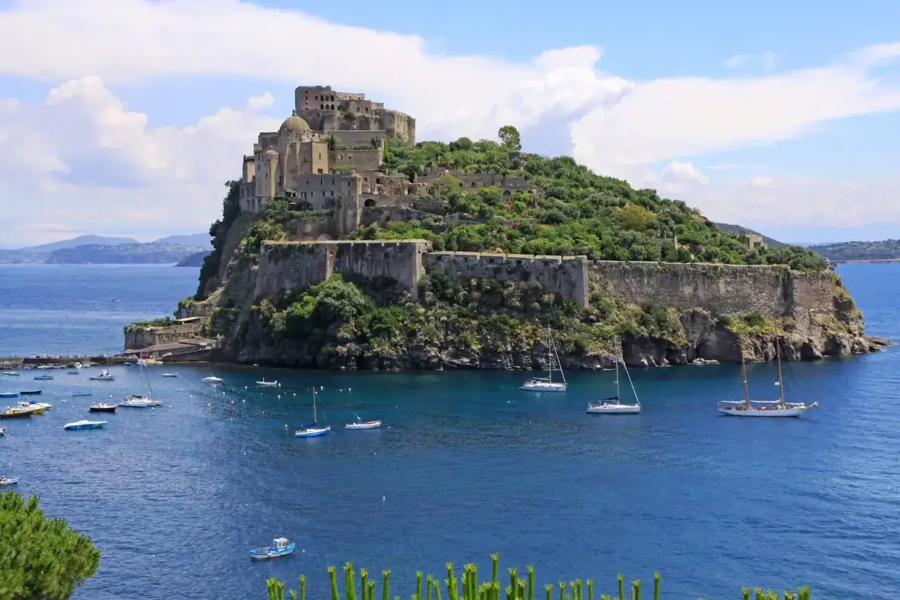 Castello Aragonese su un'isola verdeggiante, con barche a vela nel mare blu. Per sapere come arrivare ad Ischia in treno, si può raggiungere Napoli e poi prendere un traghetto.
