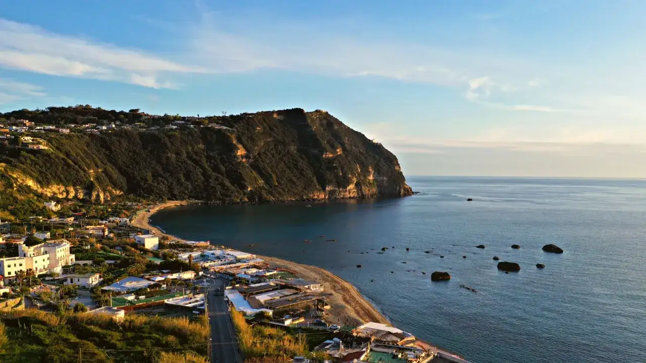 Ischia ad agosto: scorcio di costa con case bianche, spiaggia e mare blu sotto un cielo sereno.