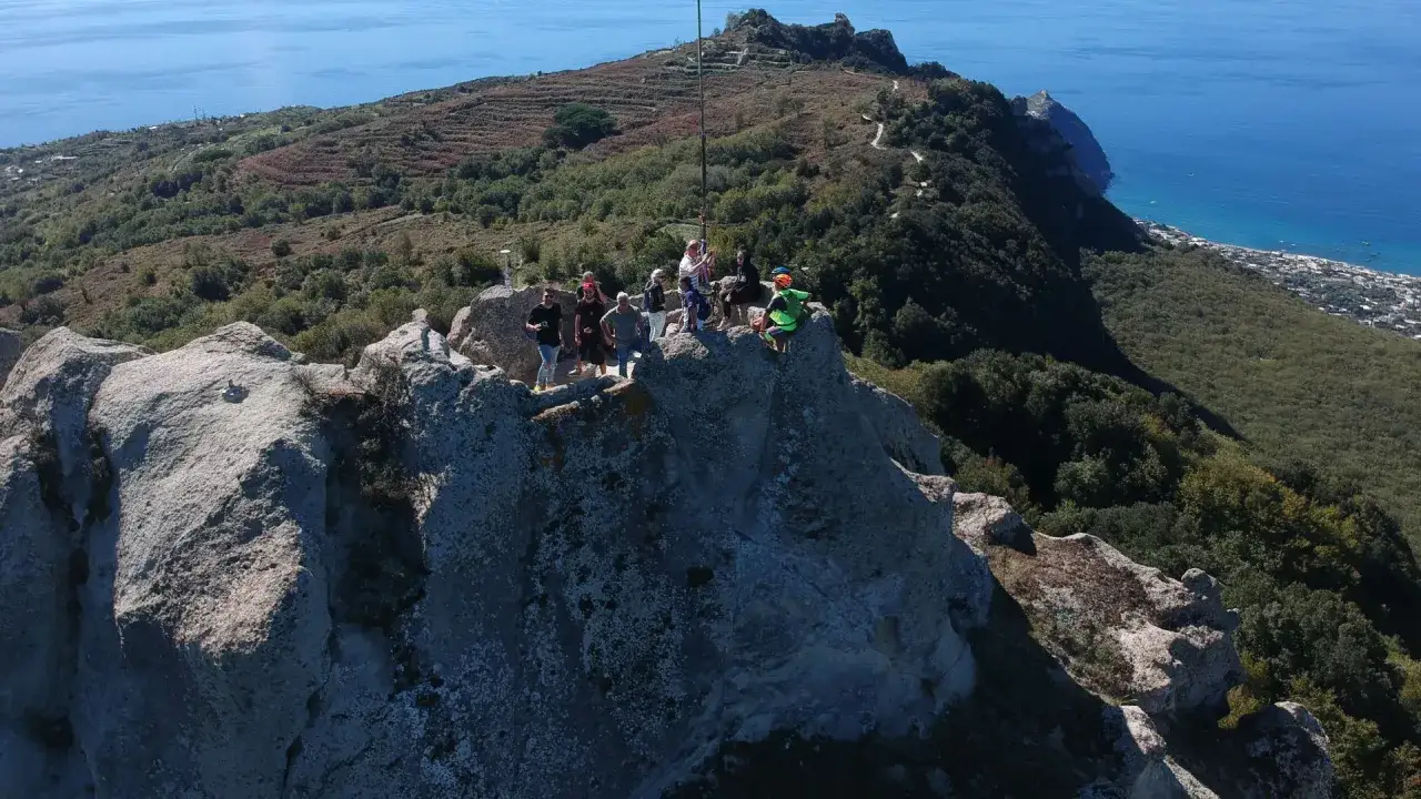 Un gruppo di persone ammira il panorama dalla cima del monte Epomeo, con il mare blu sullo sfondo.