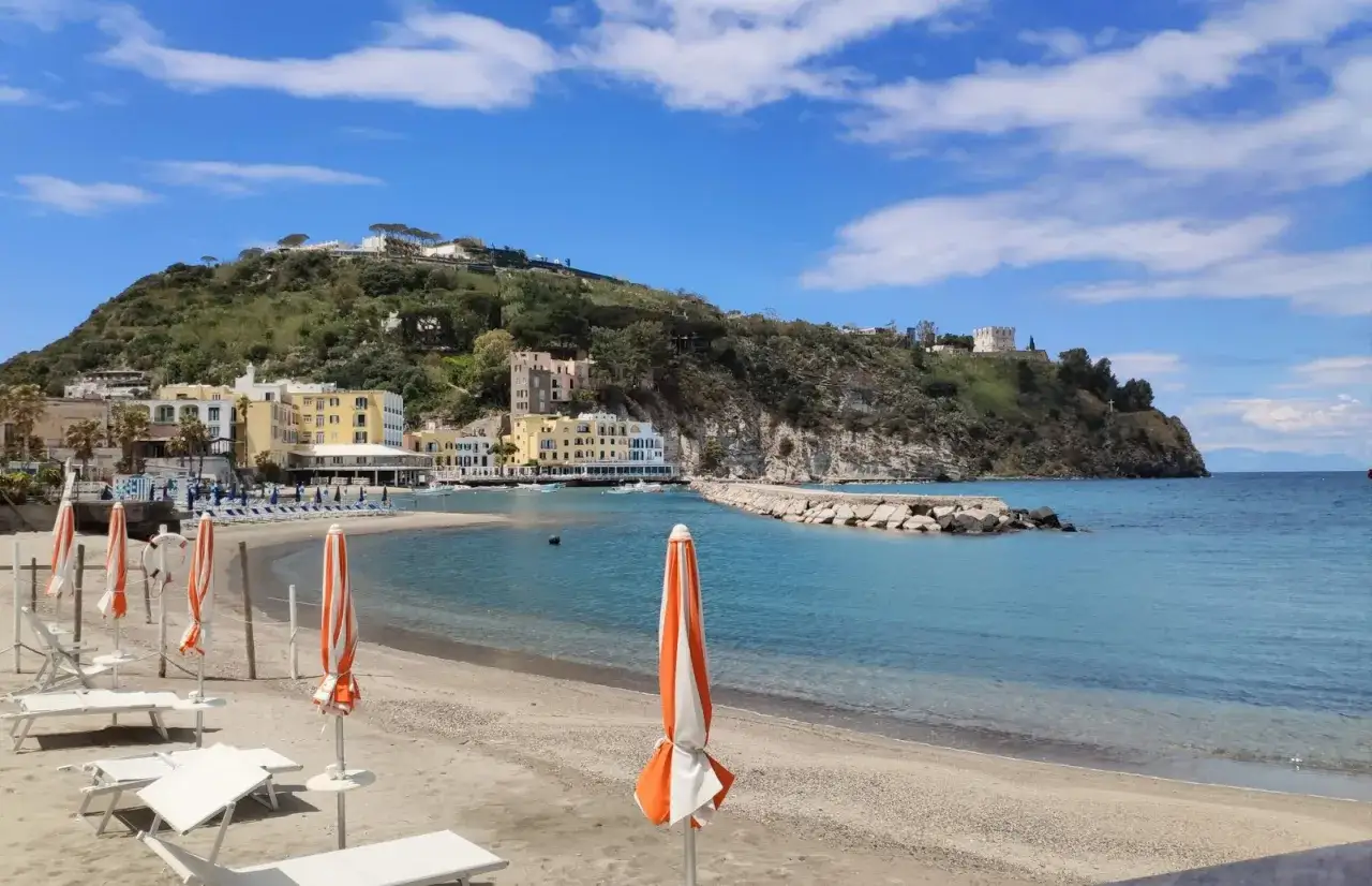 Spiaggia di Fortuna a Ischia, con lettini e ombrelloni pronti per accogliere i bagnanti. Il mare azzurro lambisce la sabbia dorata, mentre sullo sfondo si stagliano colorati edifici e una collina verdeggiante.