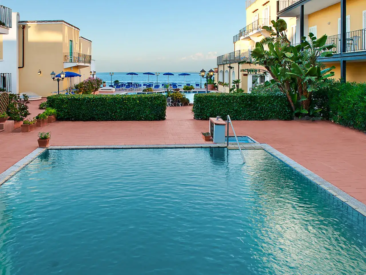 Piscina e mare al hotel Parco Aurora Terme Ischia. Ombrelloni blu e vegetazione lussureggiante invitano al relax.