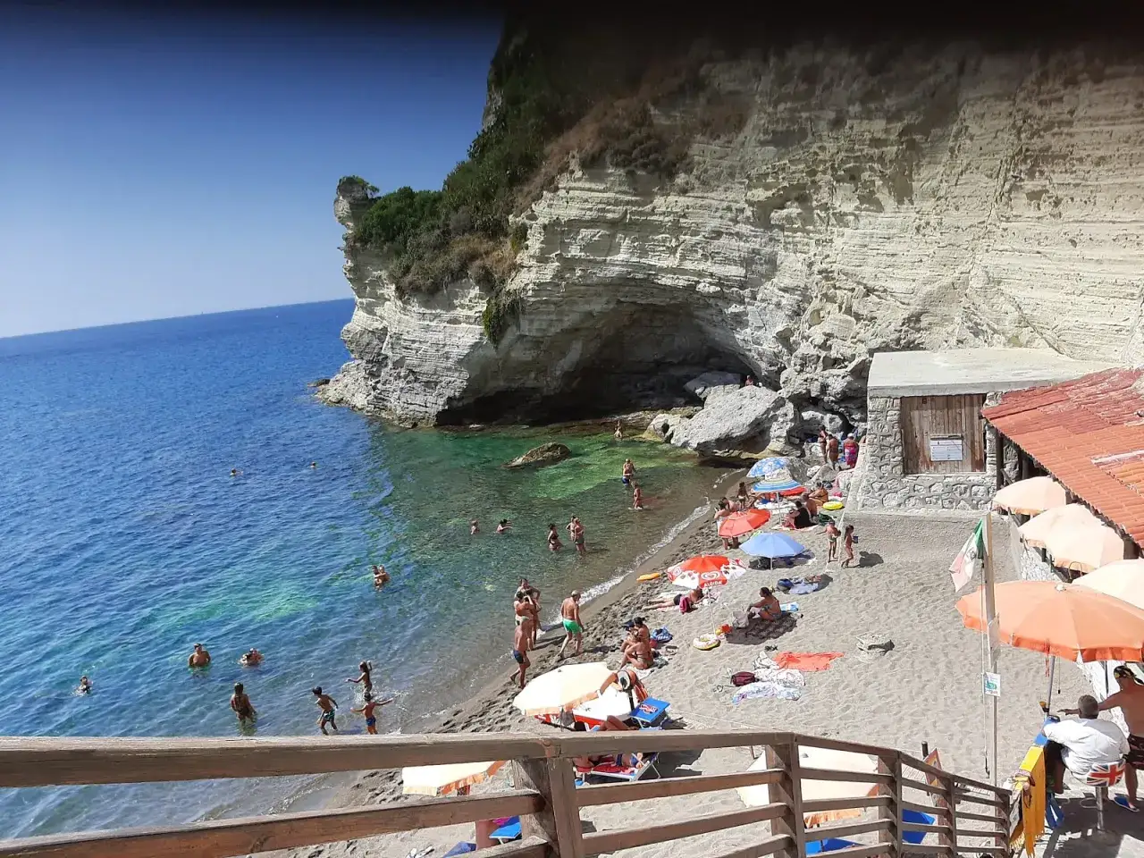 Spiaggia affollata con scogliera imponente, bagnanti nell'acqua cristallina e ombrelloni colorati. La cava grado Ischia offre un rifugio naturale.