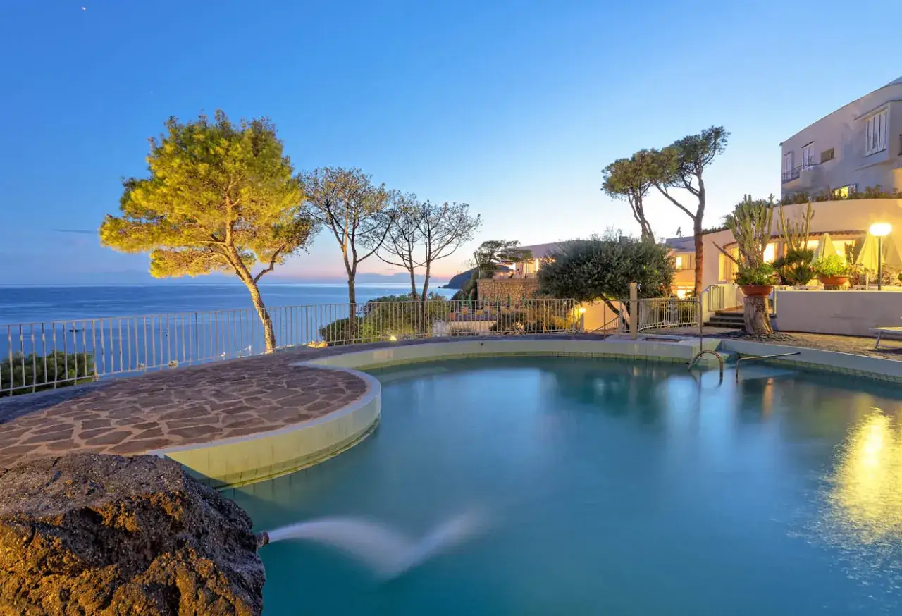 Piscina panoramica con vista mare al tramonto, alberi e la struttura del hotel oasi ischia illuminata.