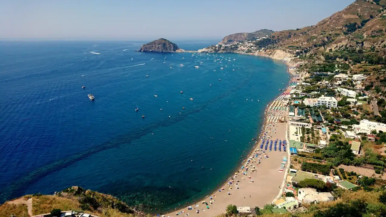 Vista aerea della spiaggia del convento, con ombrelloni colorati, mare blu intenso e barche all'orizzonte.