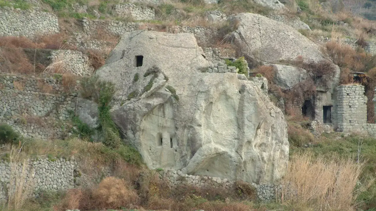 Antiche case in pietra scavate nella roccia e costruite con muretti a secco, immerse in un paesaggio collinare brullo.
