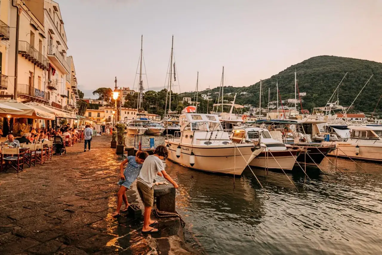 Bambini giocano sul molo della riva destra Ischia, con ristoranti affollati e barche ormeggiate al tramonto.