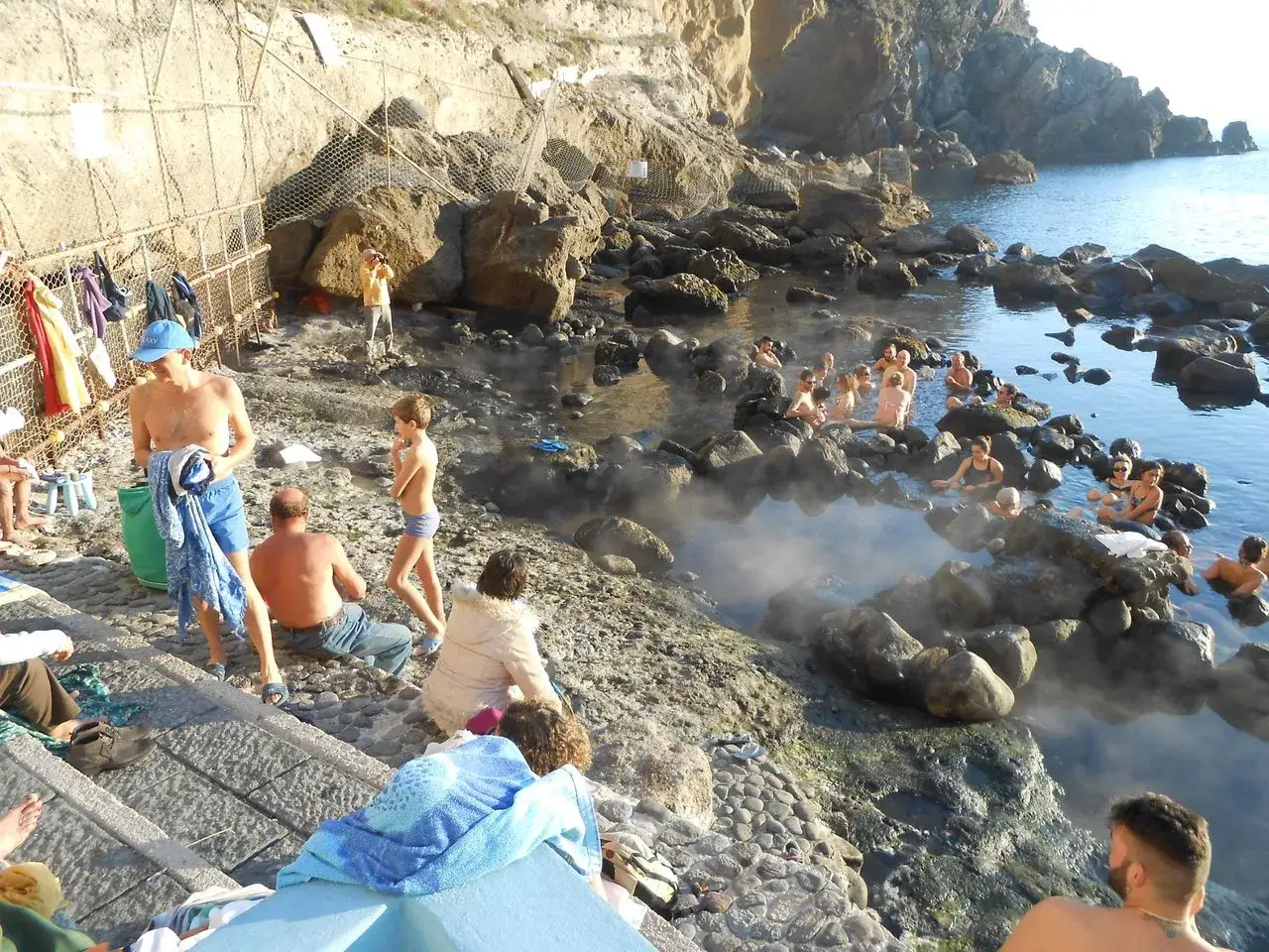 Persone si rilassano nelle acque termali della baia di Sorgeto, tra rocce e vapore.
