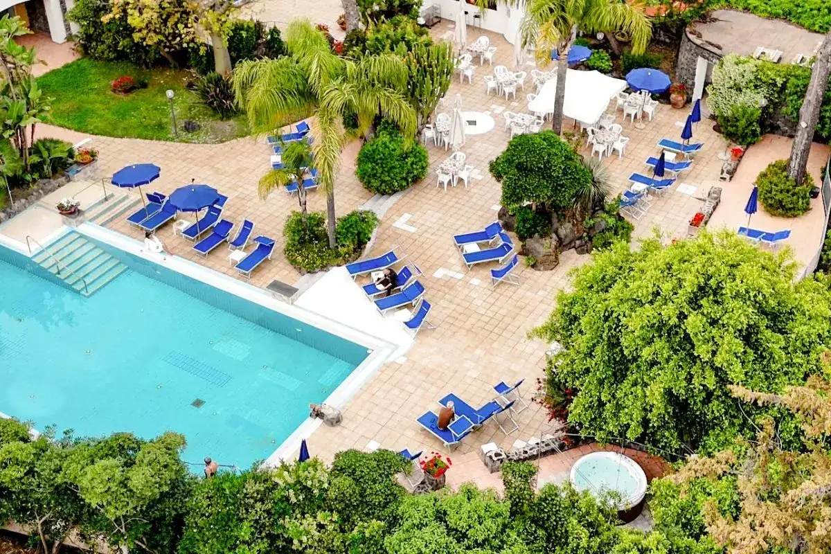 Piscina e lettini prendisole al hotel San Giovanni Ischia, circondati da vegetazione lussureggiante e un'area con tavoli all'aperto.