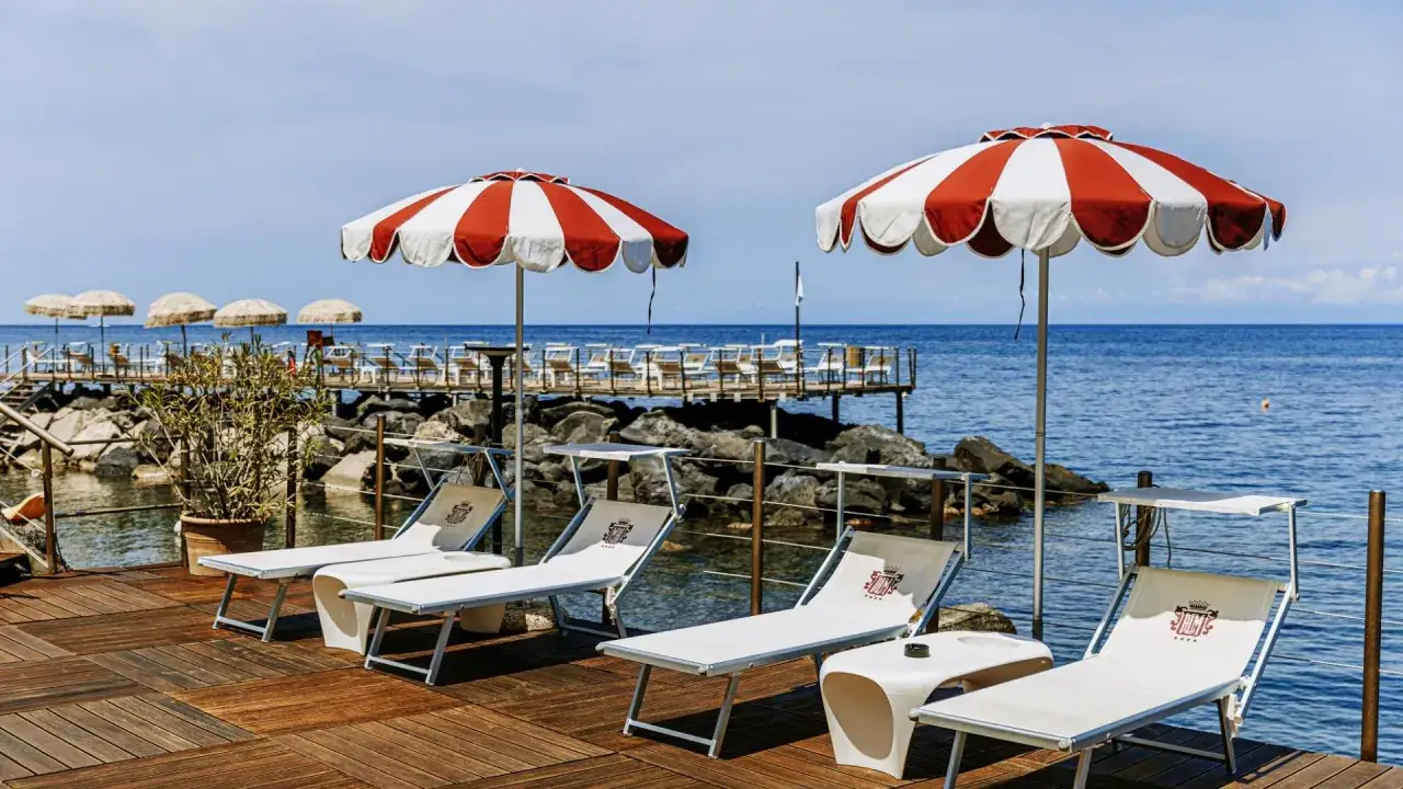 Lettini e ombrelloni a righe rosse e bianche su un pontile di legno, con il mare blu sullo sfondo. Un'oasi di relax in un beach club a Ischia.