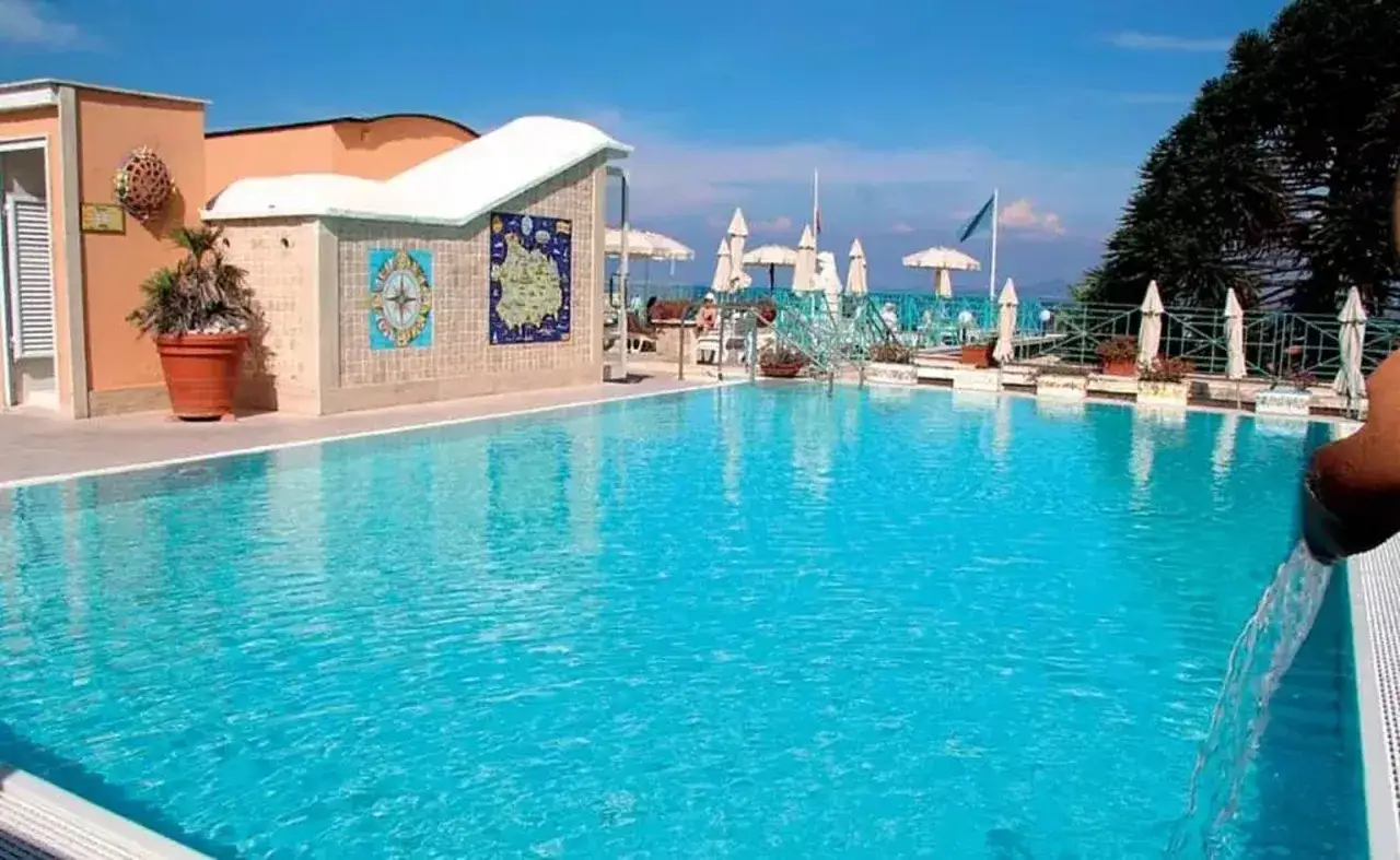 Piscina azzurra con vista mare all'Hotel Augusto Lacco Ameno. Ombrelloni bianchi e un cielo limpido.