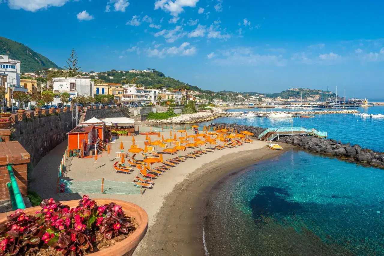 Spiaggia della Marina di Ischia con ombrelloni arancioni, lettini, mare cristallino e un porto con barche.