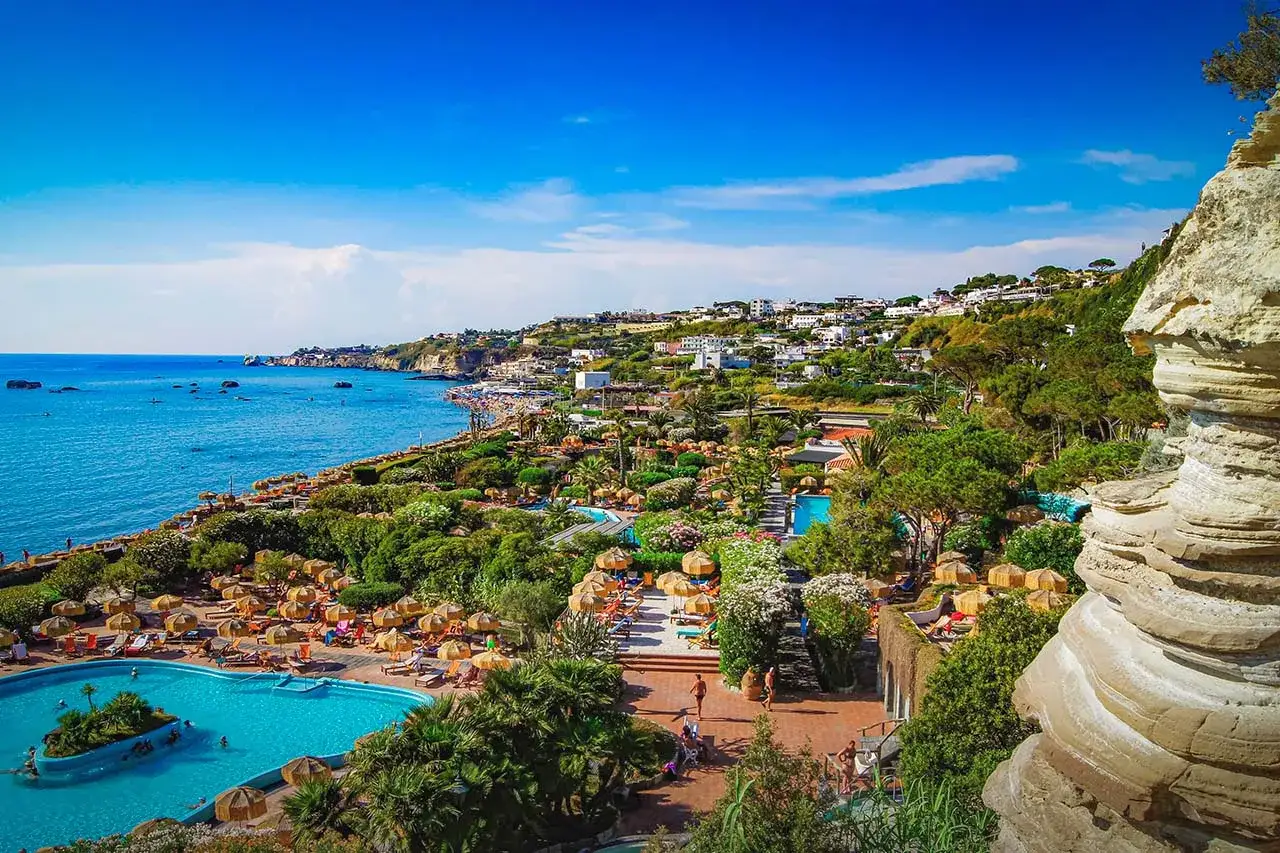 Un incantevole parco termale romantico con piscine azzurre, lettini, ombrelloni e vegetazione lussureggiante, affacciato sul mare blu e su un villaggio collinare.