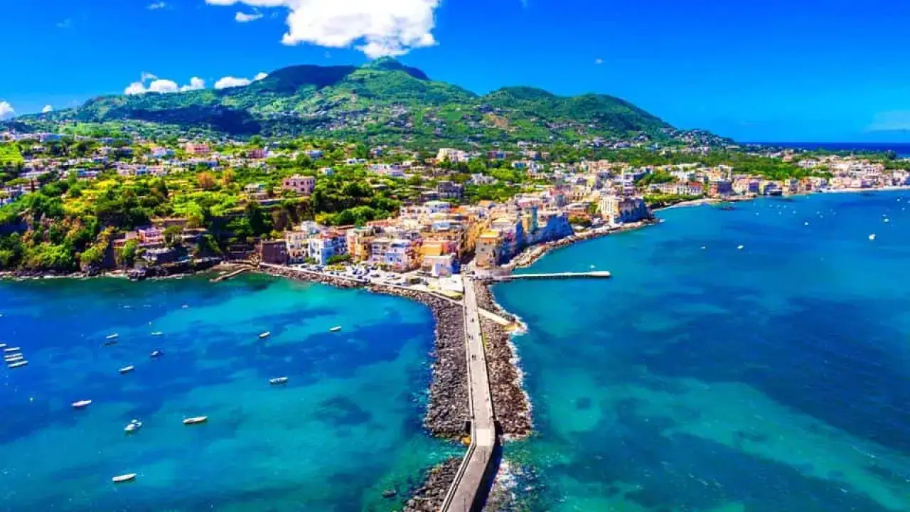 Ischia a settembre: vista aerea di un borgo colorato affacciato su un mare cristallino, con un pontile che si protende nell'acqua.