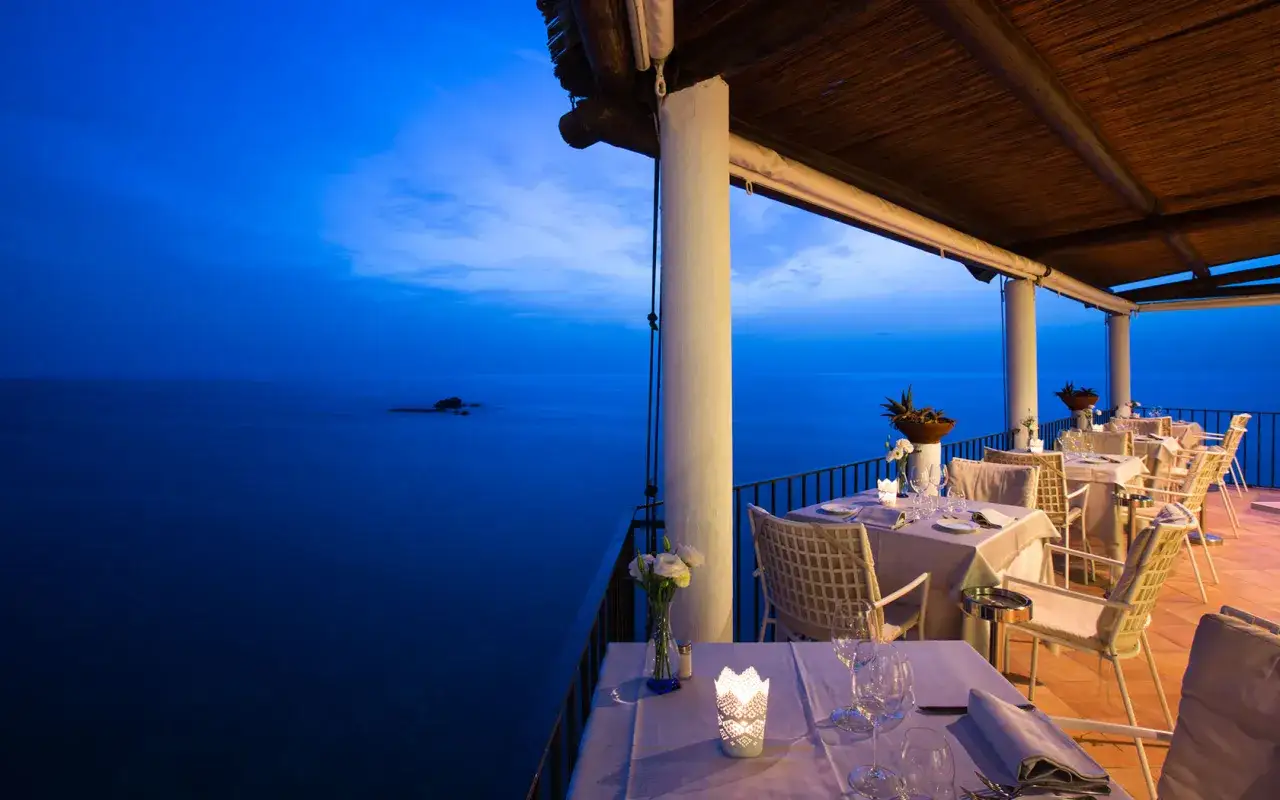 Tavoli apparecchiati su una terrazza con vista sul mare blu intenso, un invito a cena a Ischia.