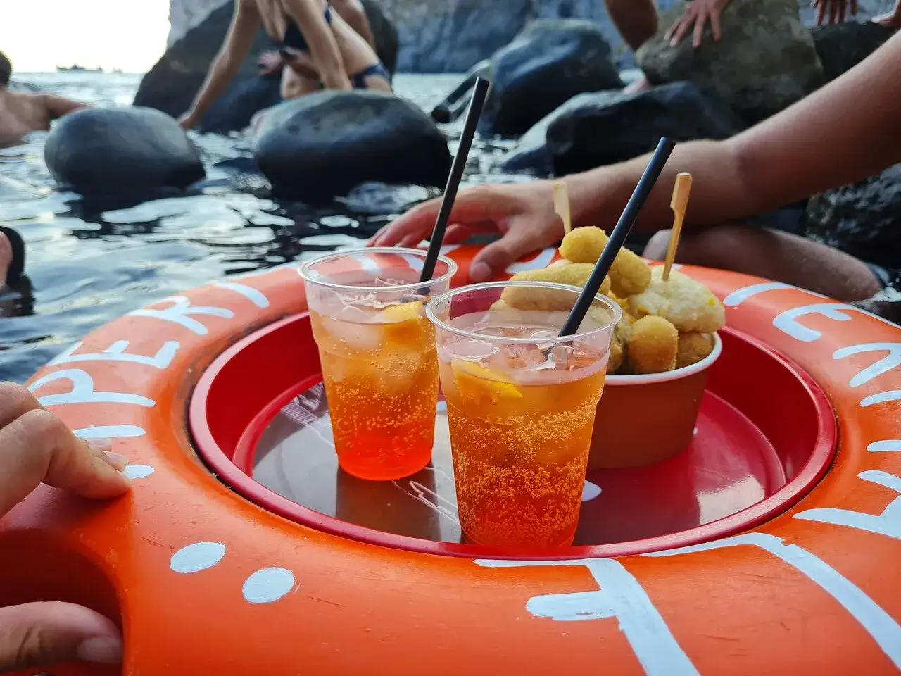 Aperitivo al sorgeto di Ischia: cocktail arancioni e frittini su un vassoio galleggiante, con persone che si godono il mare e le rocce sullo sfondo.