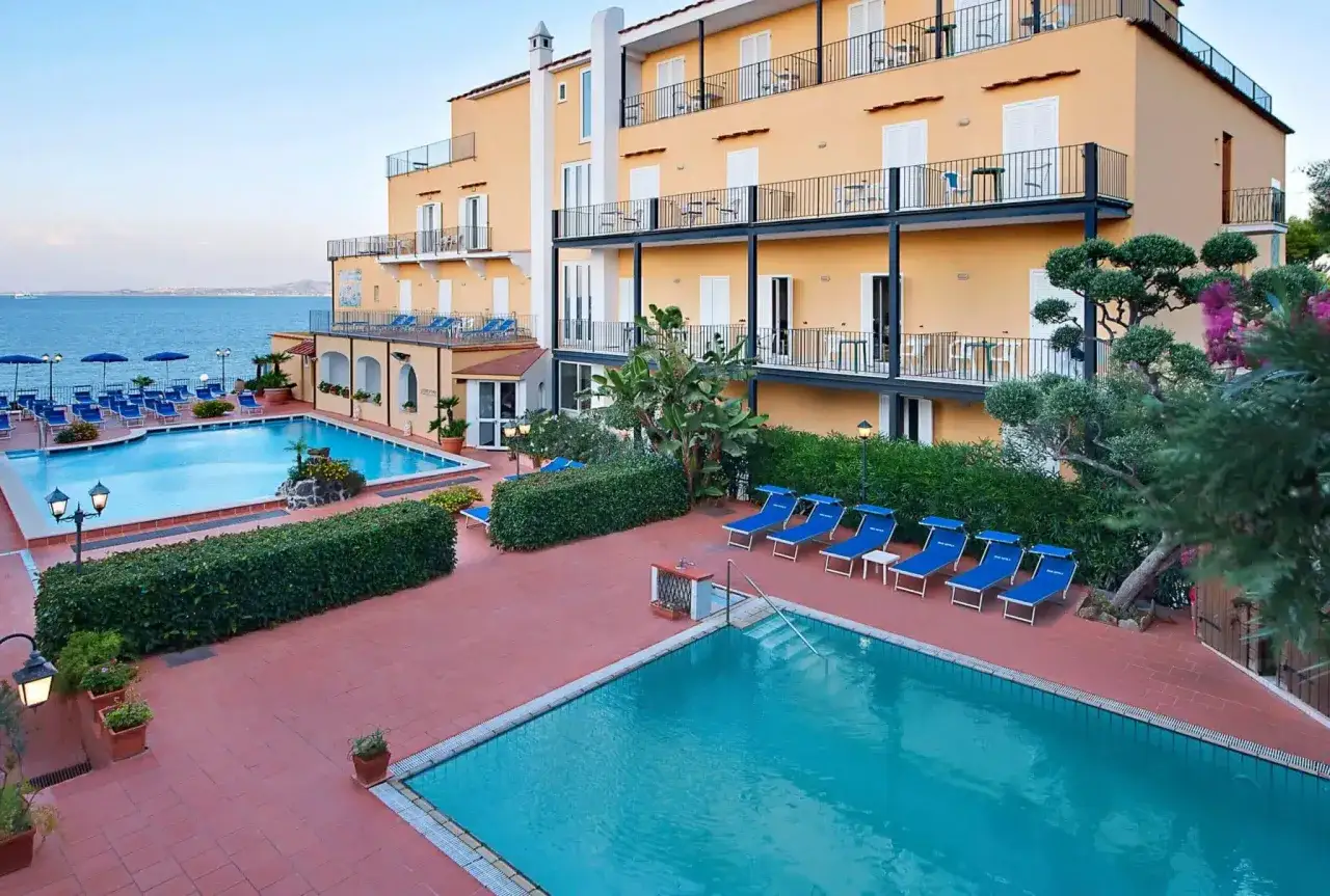 Piscine e lettini blu al Hotel Parco Aurora Terme Ischia, con vista sul mare e un edificio giallo con balconi.