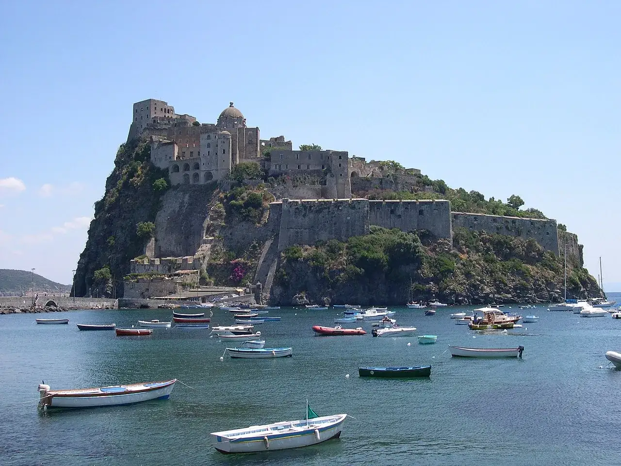 Castello Aragonese su un isolotto roccioso, con barche nel mare. Gli abitanti di Ischia sono ischitani.