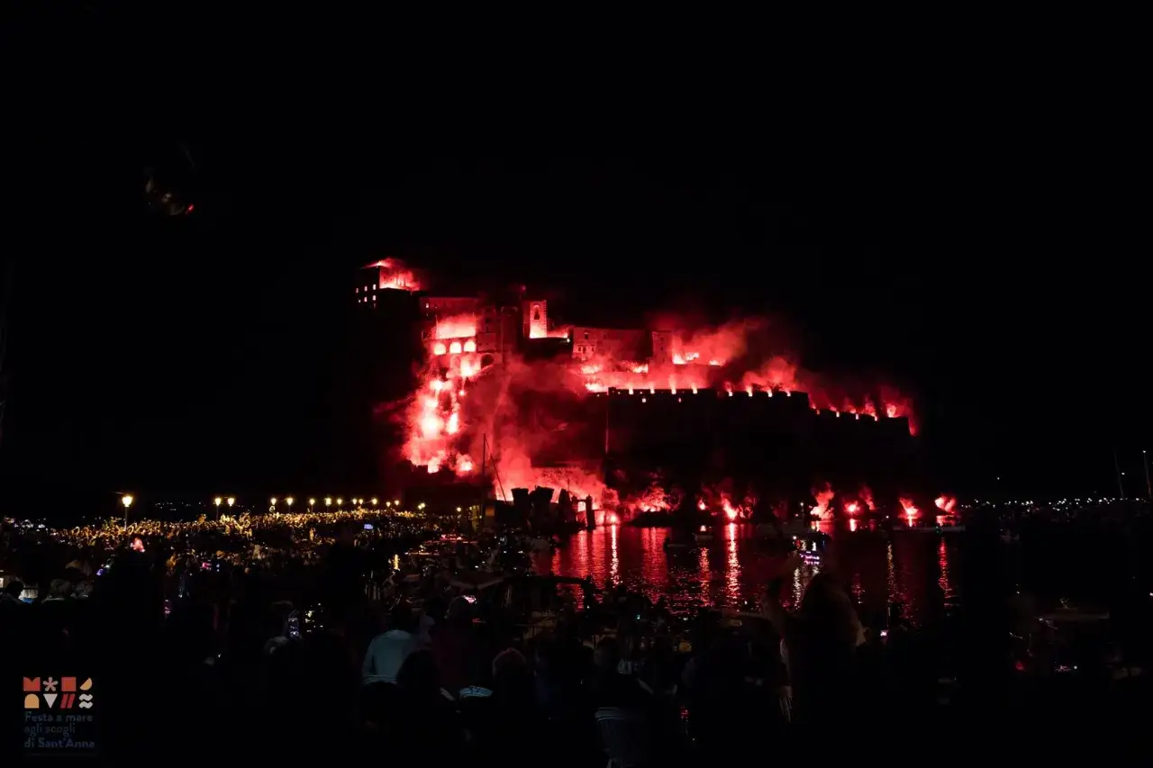 Spettacolare festa a Ischia con il castello illuminato da fuochi rossi e una folla che assiste dallo specchio d'acqua.