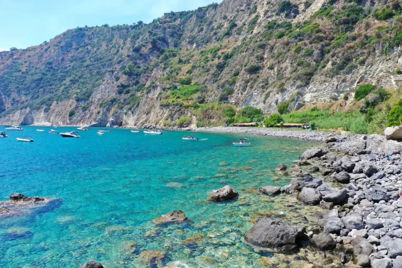 La scarrupata a Ischia: acque cristalline, scogli e barche ormeggiate sotto una costa rocciosa e verdeggiante.