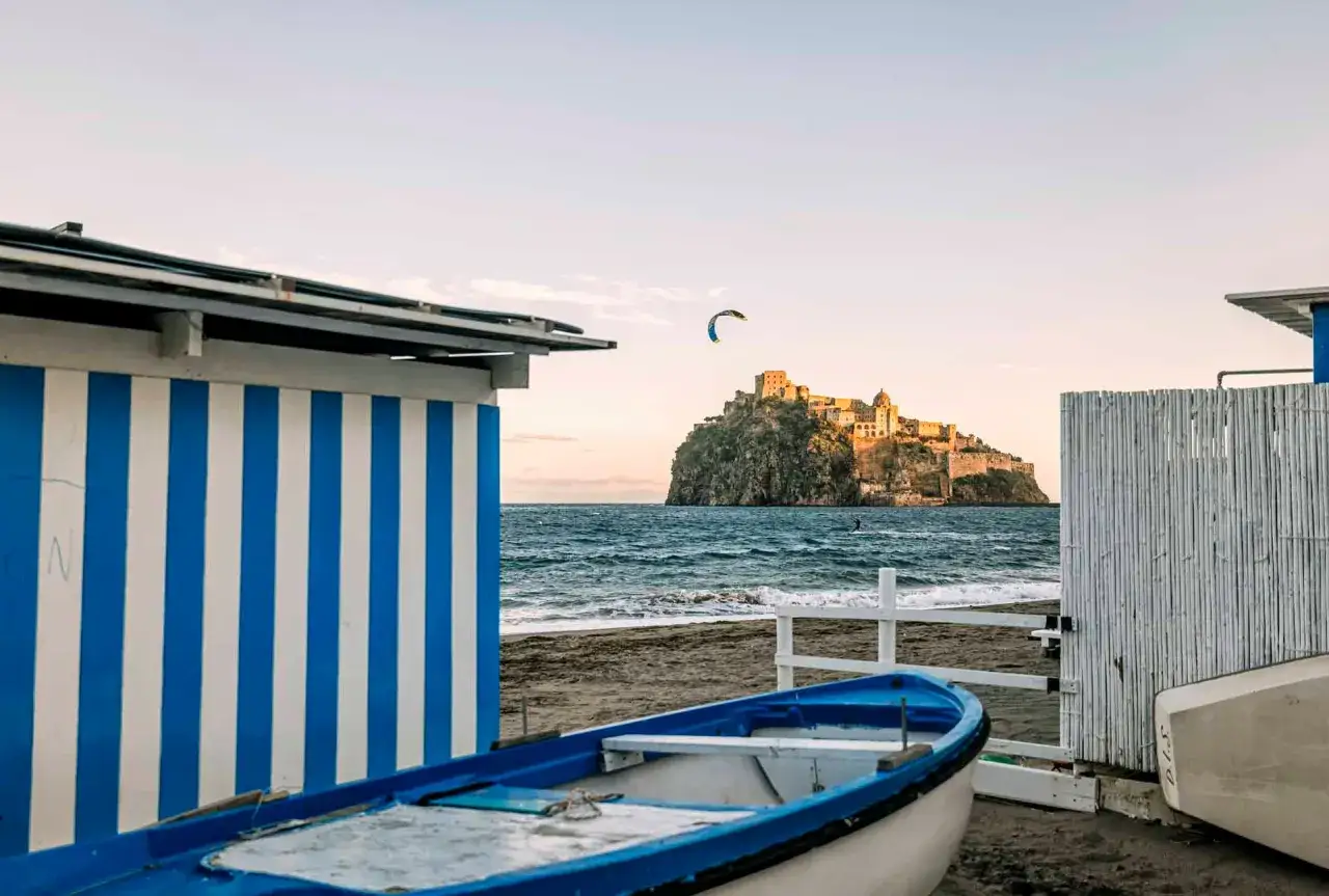 Cabine a righe blu e bianche sulla spiaggia della Mandra, Ischia, con un castello su uno scoglio in lontananza e un kitesurfer.