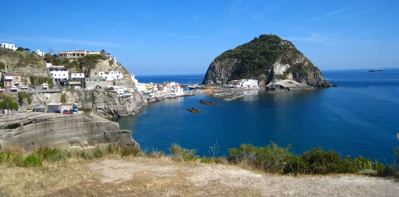 Vista panoramica di un borgo costiero con case bianche aggrappate alla roccia e un porto con barche. L'hotel Apollon si affaccia sul mare blu intenso.